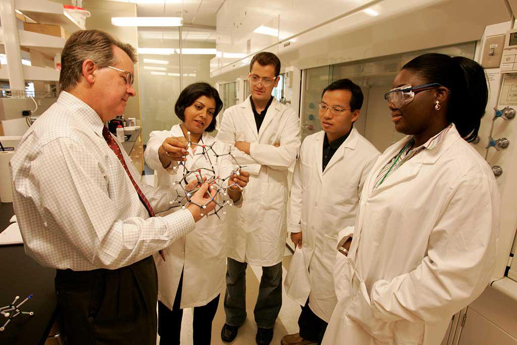 R1状态. 五名研究人员，大多穿着实验服，戴着眼镜，正在讨论一个由两名研究人员拿着的3D模型.