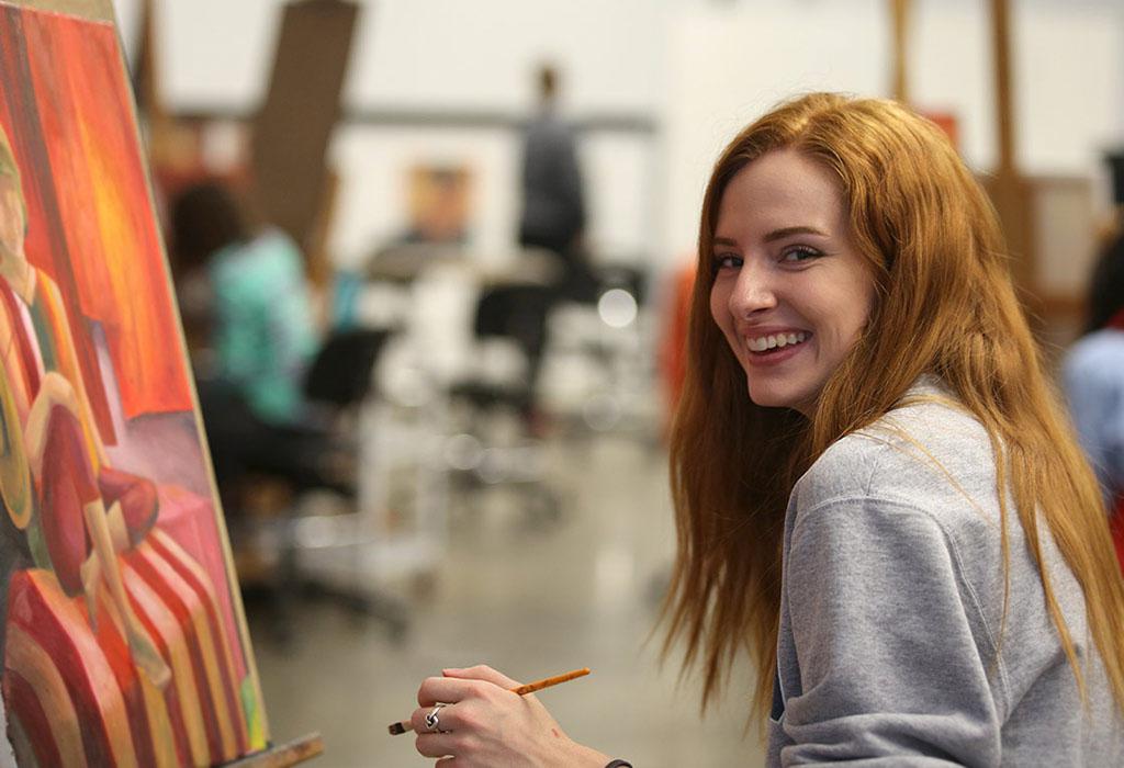 一个学生坐在一幅画前，手里拿着画笔. 他们暂停了工作，对着镜头微笑. 